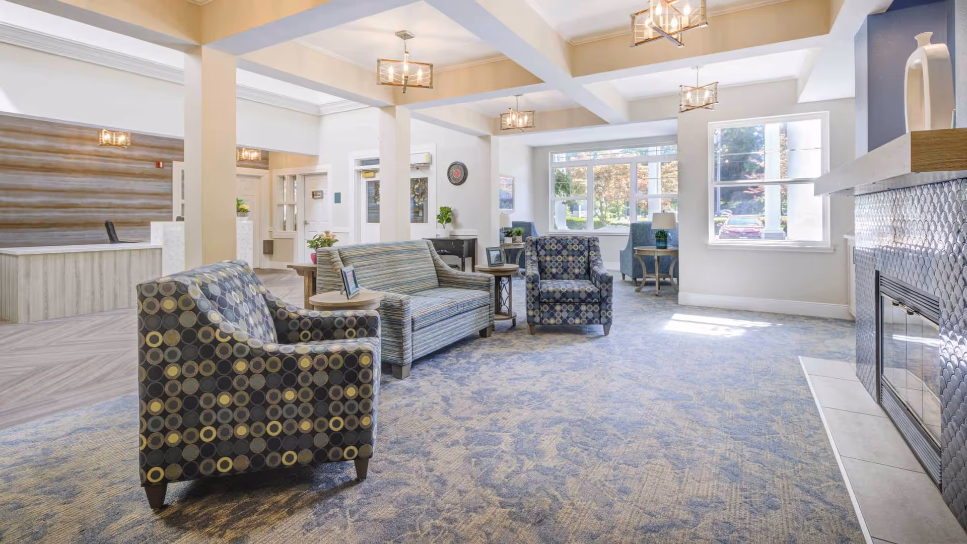 Bright furnished assisted living common room with patterned armchairs, a sofa, large windows, and a fireplace.