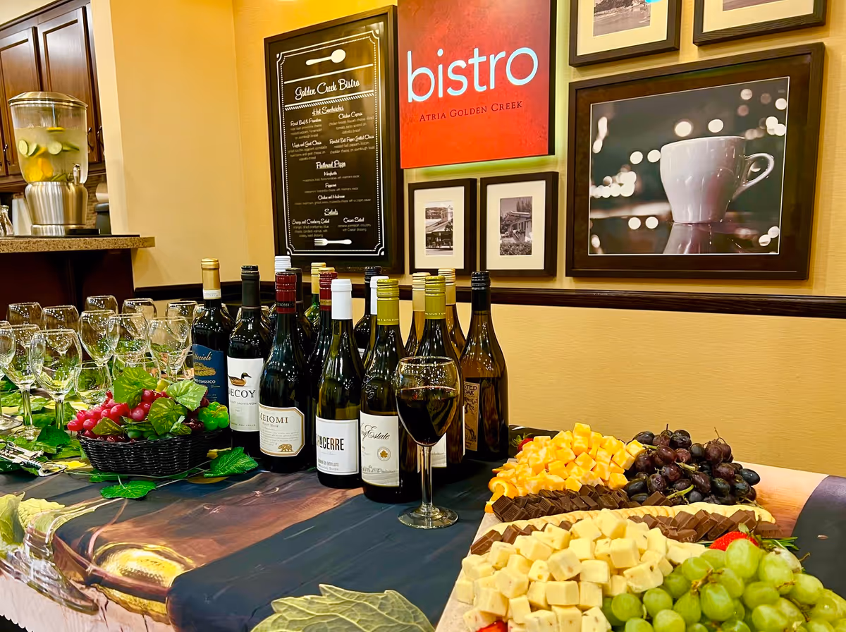 A table set with multiple bottles of wine, wine glasses, a basket of grapes, and a platter of assorted cheeses and grapes. Behind the table, there are framed pictures and a sign that reads 'bistro Atria Golden Creek' along with a menu board titled 'Golden Creek Bistro'.