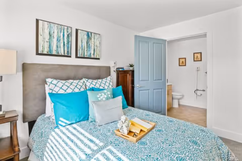 A bedroom with a bed featuring a blue and white patterned bedspread and multiple pillows in shades of blue and gray. Above the bed are two abstract paintings with blue and green tones. To the right, an open door reveals a bathroom with a toilet and two framed pictures on the wall. A wooden nightstand with a lamp is visible on the left side of the bed.