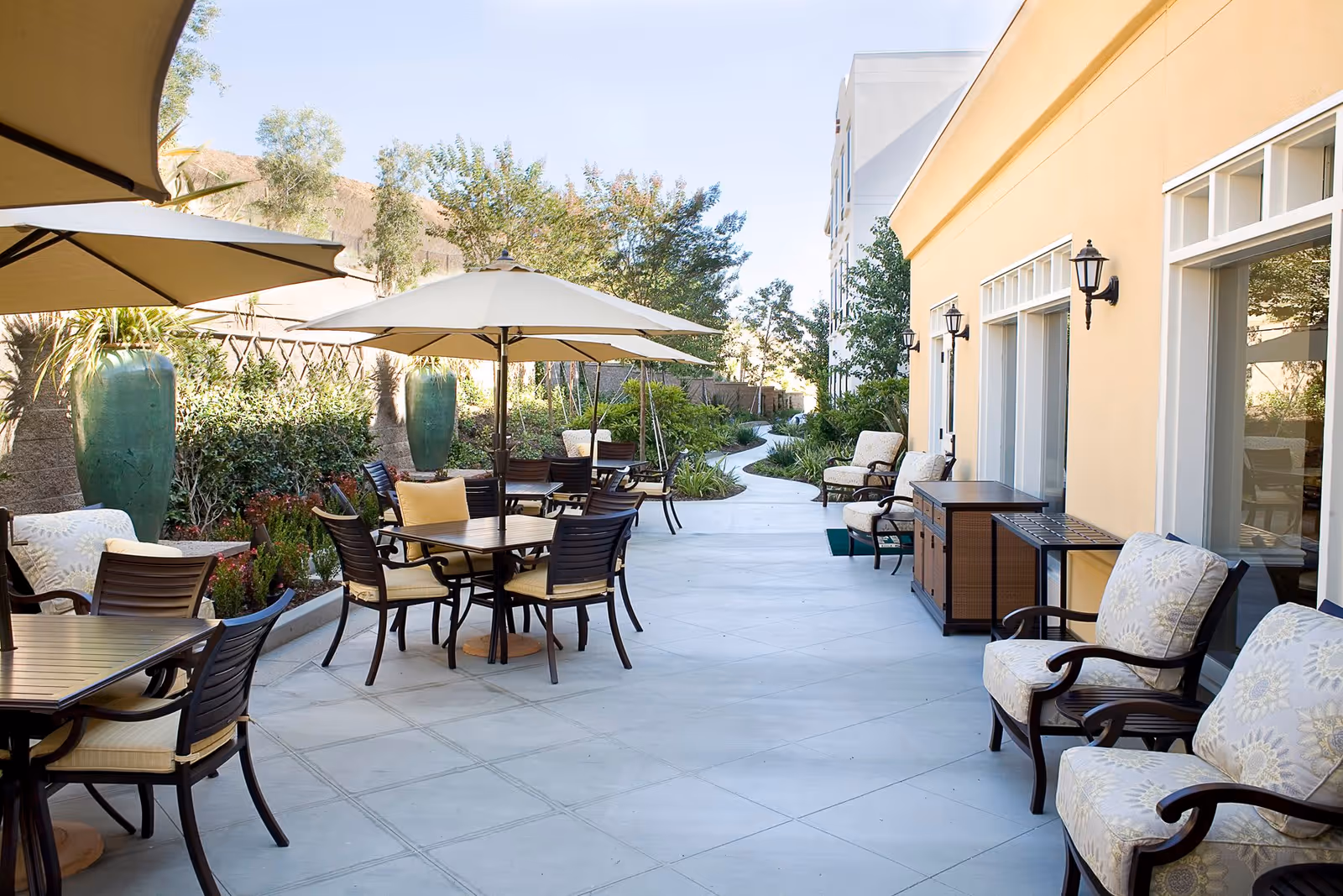 Outdoor patio area at Belmont Village Thousand Oaks with multiple tables and chairs under large beige umbrellas, cushioned armchairs along the building wall, large green decorative vases, and a winding pathway surrounded by greenery and trees.