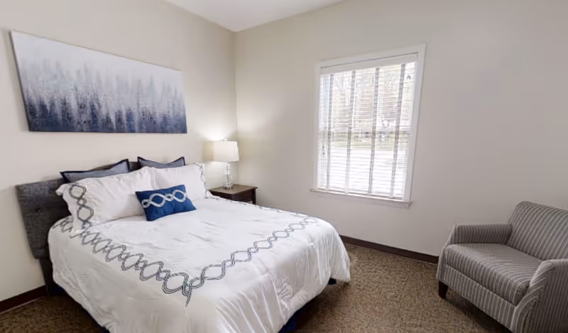 A neatly made bed with white and blue bedding in a simple bedroom. There is a nightstand with a lamp next to the bed, a window with blinds letting in natural light, a gray upholstered armchair, and a piece of abstract artwork hanging above the bed.