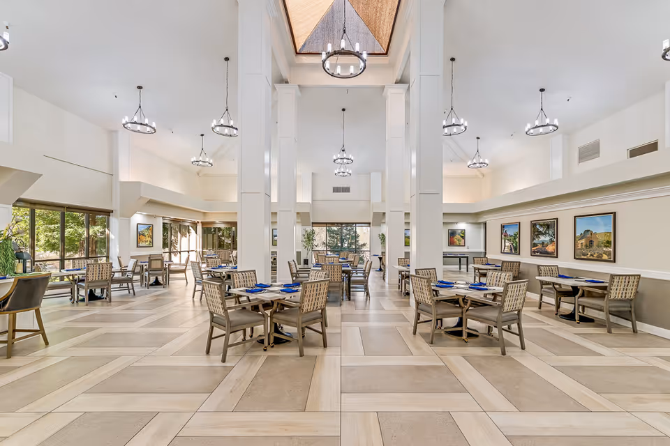 Bright, spacious dining room with multiple tables and chairs, high ceilings with chandeliers, large windows and framed artwork.