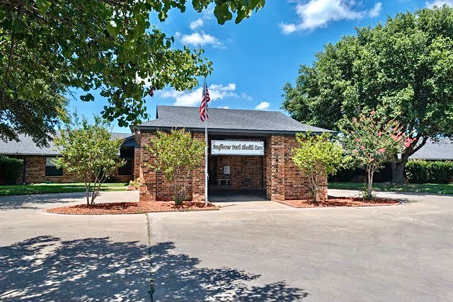 Front exterior view of Sunflower Park Health Care building with a brick facade, an American flag on a flagpole, and trees on either side under a blue sky with some clouds.