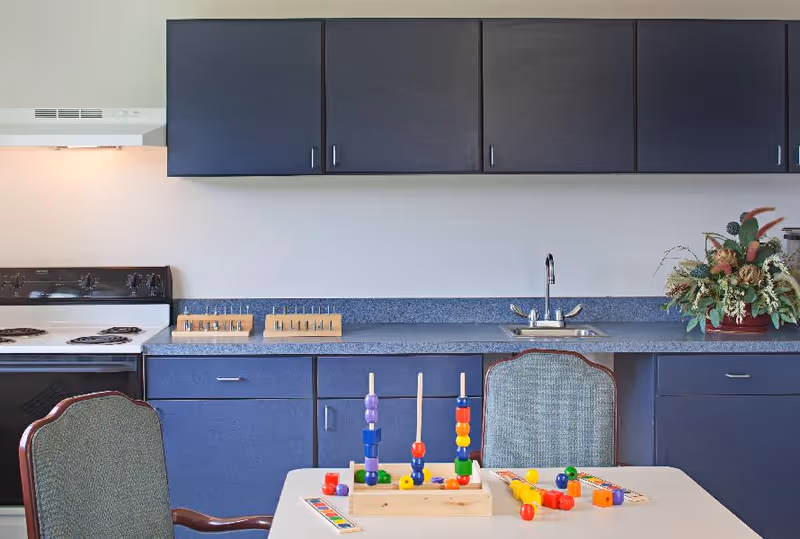 A kitchen area with dark blue cabinets and a countertop featuring a sink and a stove. In front of the counter is a table with colorful wooden bead toys and two chairs with green upholstery. There is a floral arrangement on the right side of the counter.