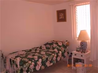A bedroom with a single bed, floral quilt, nightstand, lamp, and a window.