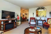 Living room with a wall-mounted TV above a fireplace, upholstered chairs, a coffee table, and a view into a dining area.
