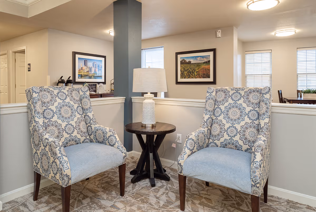 Two patterned armchairs with blue and beige floral designs are positioned on either side of a small round wooden table with a white textured lamp on it. The seating area is set against a half wall with a blue pillar in the center. Behind the seating area, there are framed pictures on the walls and windows with blinds, and a dining table with chairs is visible in the background.