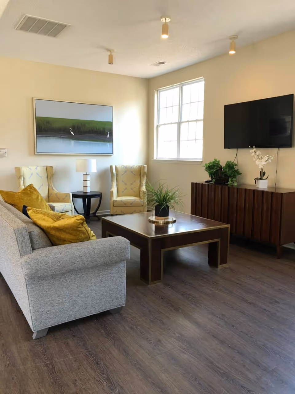 A cozy living room with a gray sofa adorned with mustard yellow pillows, two yellow patterned armchairs, a dark wooden coffee table with a small plant centerpiece, a side table with a lamp, a large window letting in natural light, a wall-mounted flat-screen TV, and a wooden cabinet with decorative plants and flowers.