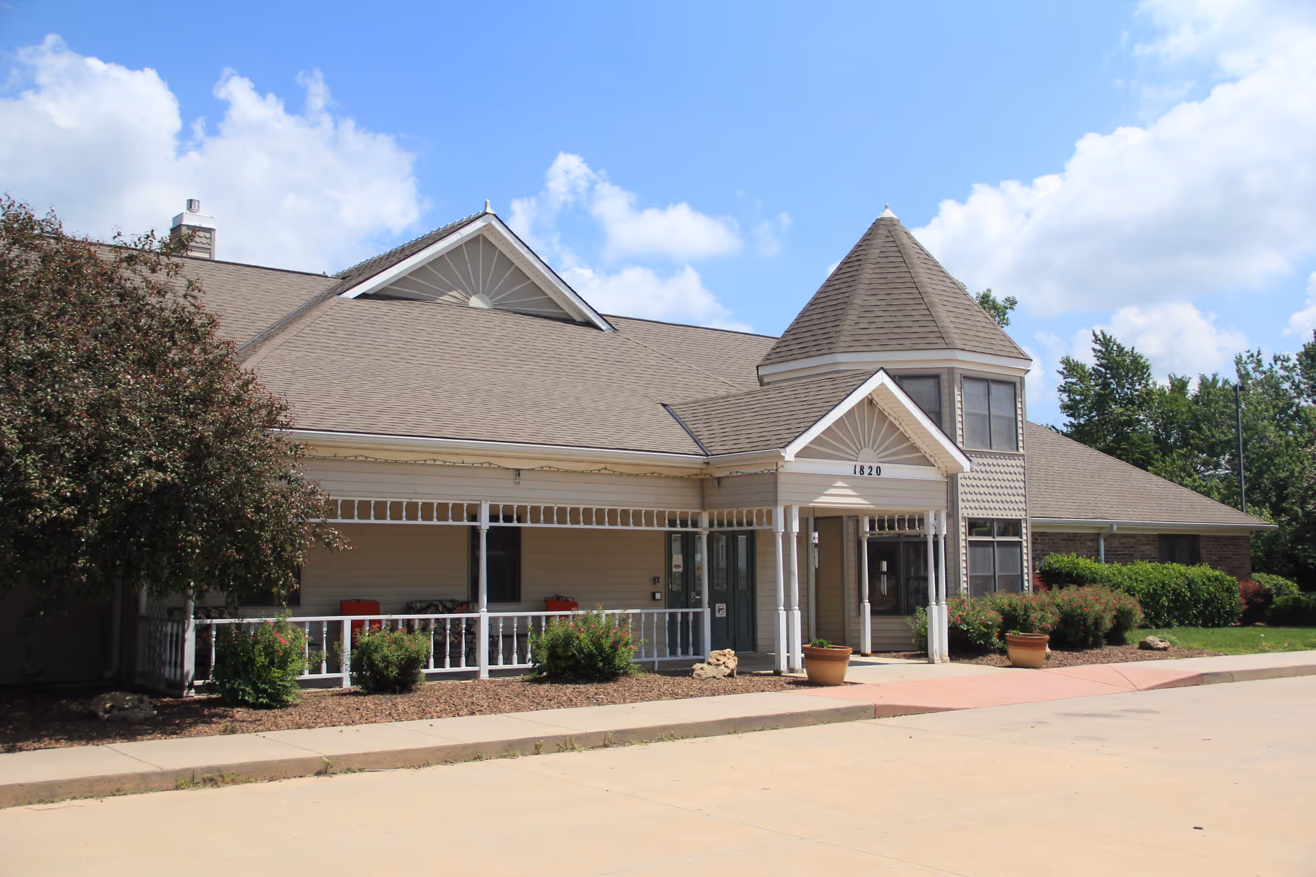 Exterior view of a single-story building with beige siding and a gray shingled roof under a blue sky with scattered clouds. The building features a covered porch with white railings and columns, a turret-like structure on the right side, and the number 1820 displayed above the entrance. There are shrubs and potted plants around the building.