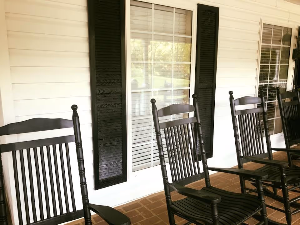 A row of black wooden rocking chairs lined up on a covered porch with white siding and windows with black shutters in the background.