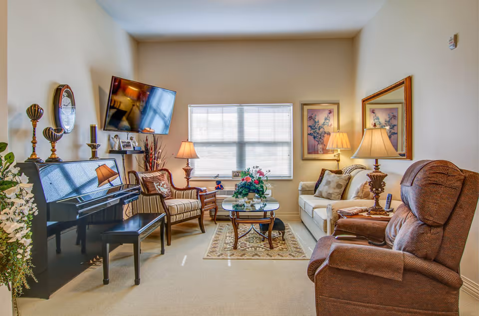 Bright living room with a sofa, recliner, armchairs, a piano, wall-mounted TV and a central coffee table with flowers.