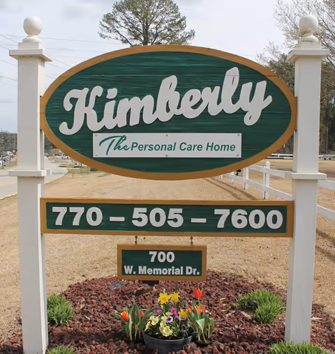 Green and white wooden roadside sign for Kimberly Personal Care Home displaying a phone number and address in a landscaped bed.