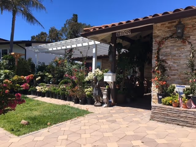 Front exterior view of a single-story building with a tiled roof and stone facade. The entrance area is decorated with numerous potted plants and flowers arranged along the walkway and under a white pergola. There is a mailbox and garden statues near the entrance. The sky is clear and blue, and a palm tree is visible on the left side.