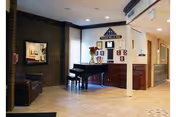 Interior view of a senior living facility common area featuring a black grand piano with a bench, a brown leather armchair, a large mirror on a brick wall, and a wooden cabinet with decorative items including a vase with flowers. The area is well-lit with recessed ceiling lights and has a tiled floor.