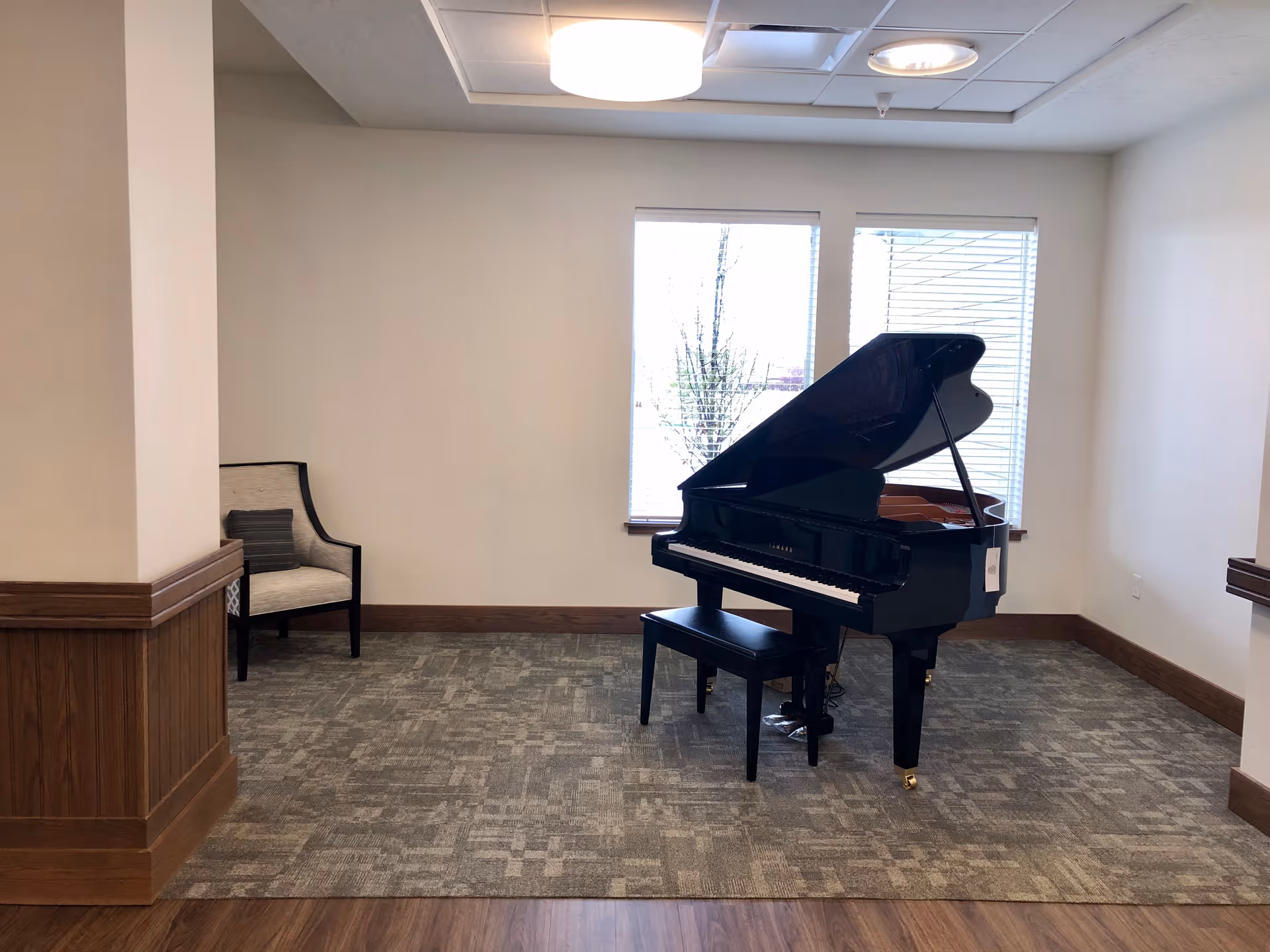 A small common room with a black grand piano and bench by two windows, a chair to the left, and neutral walls and carpeting.