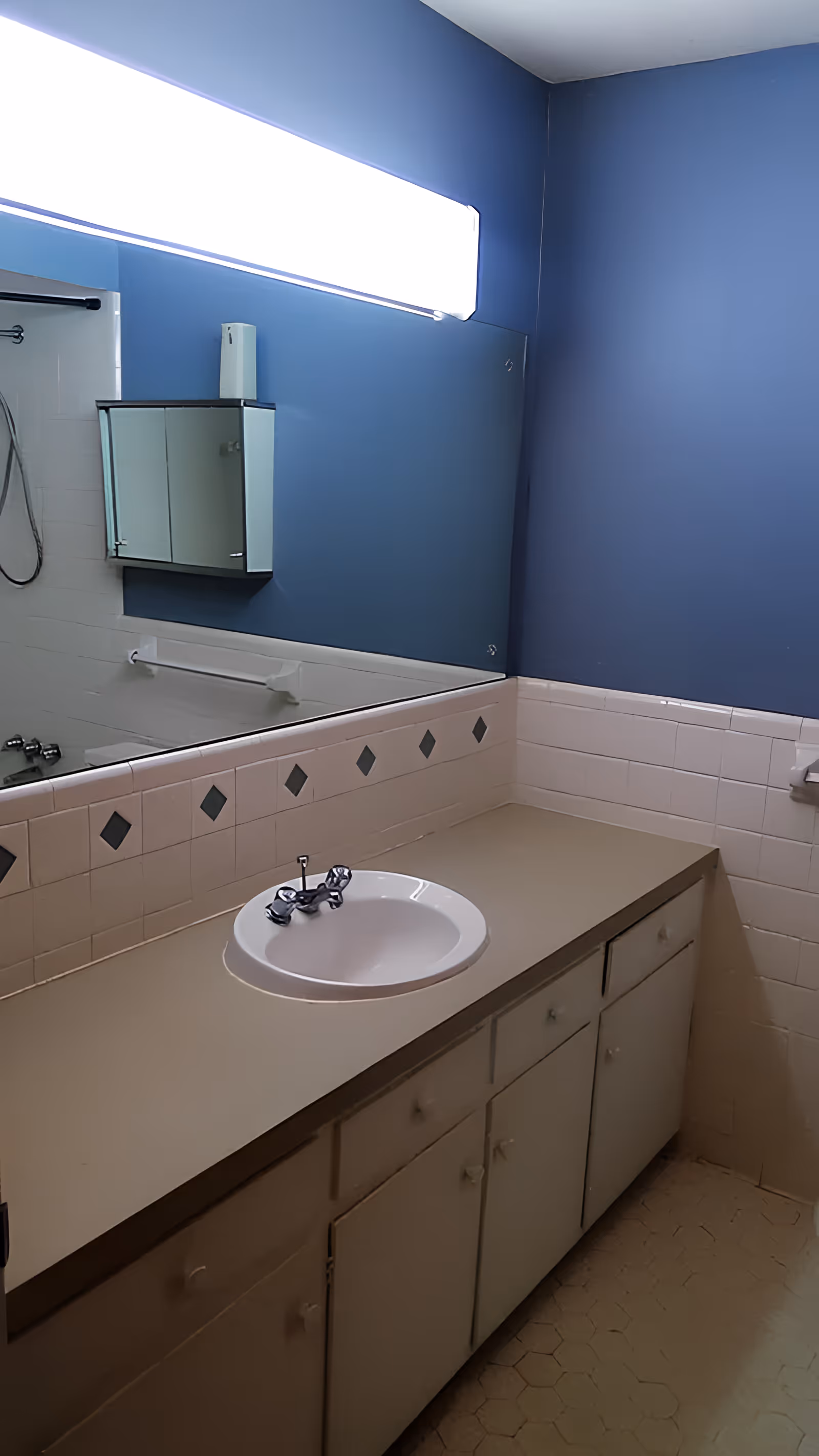 Bathroom interior with a beige countertop and white sink, a large mirror above the sink, blue walls, white tiled backsplash with diamond-shaped accents, a medicine cabinet with mirrored doors, and a shower curtain rod visible in the reflection.