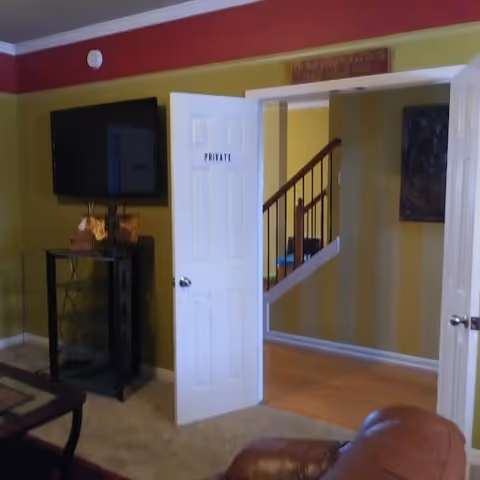 Living room with a wall-mounted TV, an open door labeled "PRIVATE" revealing a hallway and staircase.