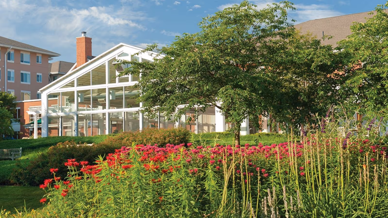 A landscaped garden with vibrant red and pink flowers, green shrubs, and a large tree in front of a building with large glass windows and a white frame. The building appears to be part of a retirement community with a clear blue sky above.