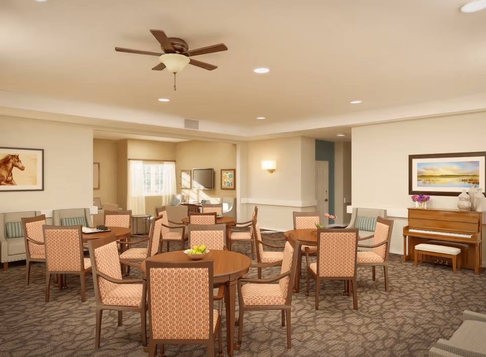 A spacious common area with multiple round wooden tables surrounded by cushioned chairs with patterned upholstery. The room features a ceiling fan, recessed lighting, a piano with a bench, framed artwork on the walls, and a carpeted floor with a leaf pattern. There are also sofas and a television in the background near a window with sheer curtains.