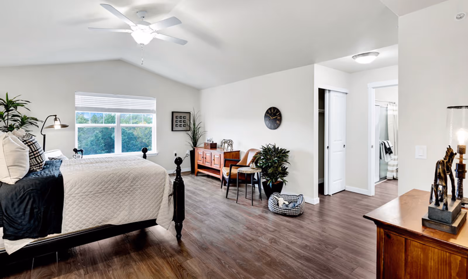 A bright and spacious bedroom with a large window showing greenery outside. The room features a bed with white and black bedding, a ceiling fan, wooden flooring, a wooden dresser with decorative items, a chair, a plant, and a pet bed. There is an open doorway leading to a bathroom with a shower visible.