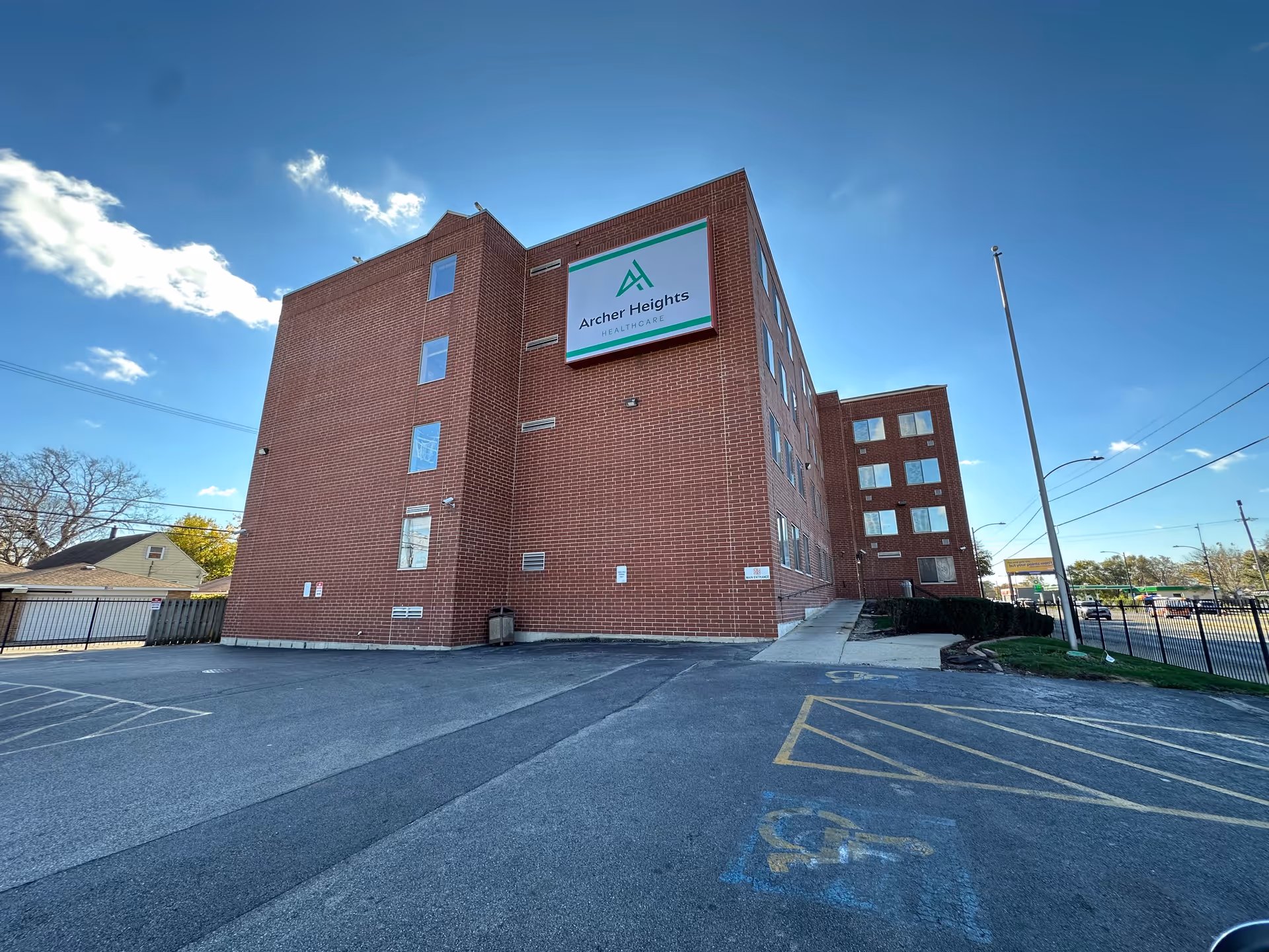 Exterior view of a multi-story brick building with a sign that reads 'Archer Heights Healthcare' on the side. The building is surrounded by a parking lot with marked handicap parking spaces and a clear blue sky with some clouds.