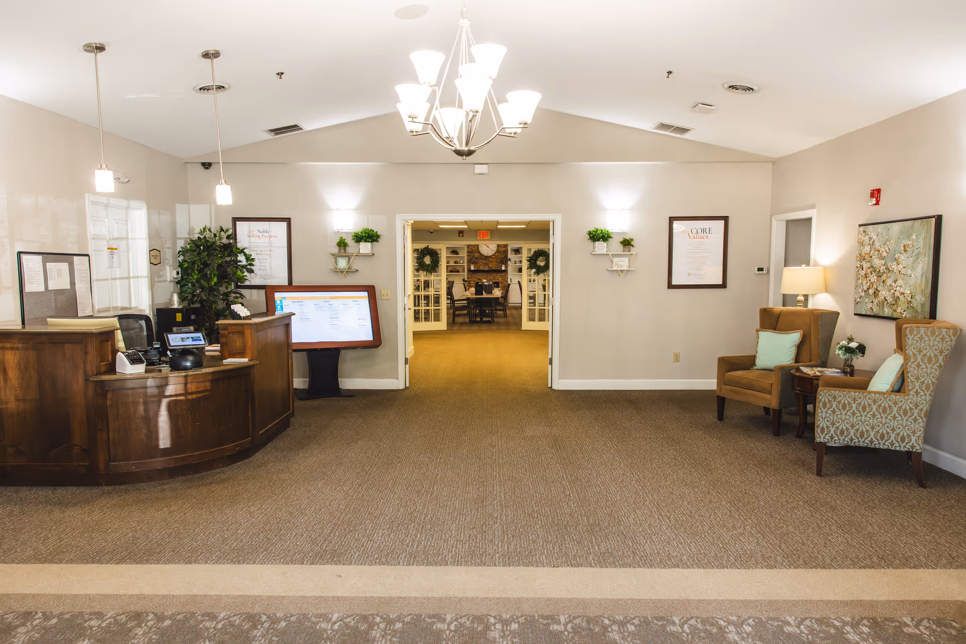 Reception area of Commonwealth Senior Living at the West End with a wooden front desk on the left, a digital information board, and two armchairs with a small table and lamp on the right. The room has beige walls, carpeted floor, and a chandelier hanging from the ceiling. Double doors in the center lead to another room with a dining table and chairs.