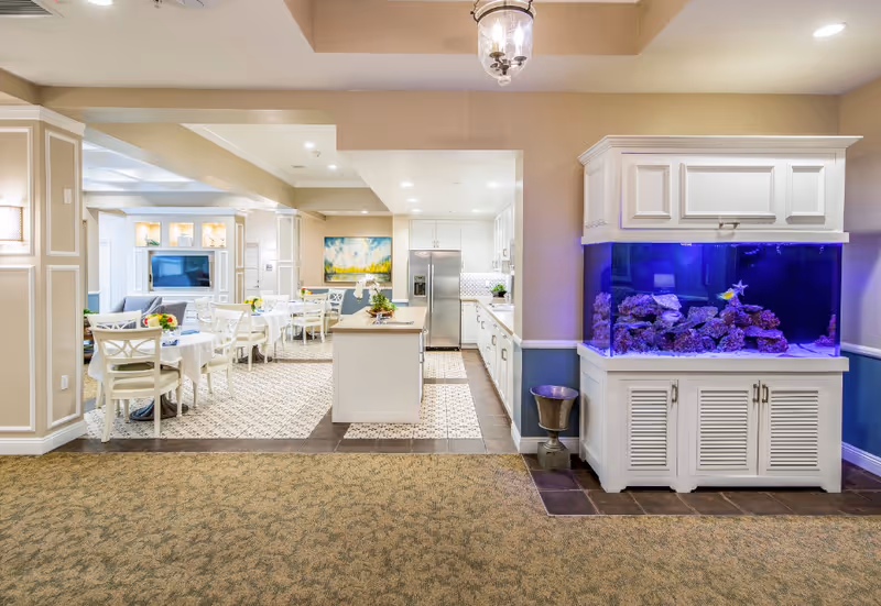 Bright communal dining and living area with white tables and chairs, a kitchen in the back, and a large aquarium on the right.