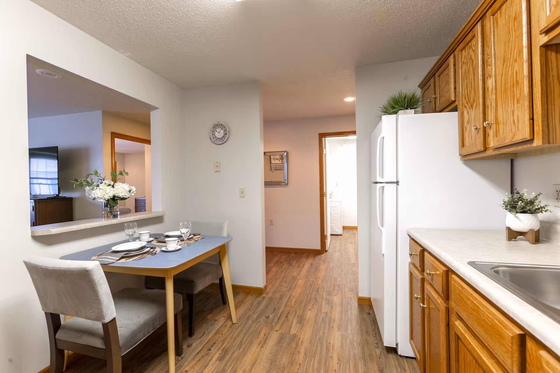 A small dining area with a table set for two, featuring plates, cups, and glasses. The dining area is adjacent to a kitchen with wooden cabinets, a white refrigerator, and a countertop with a small plant. The floor is wood, and there is a clock on the wall. A living room with a TV and a vase of flowers is visible through a pass-through window.