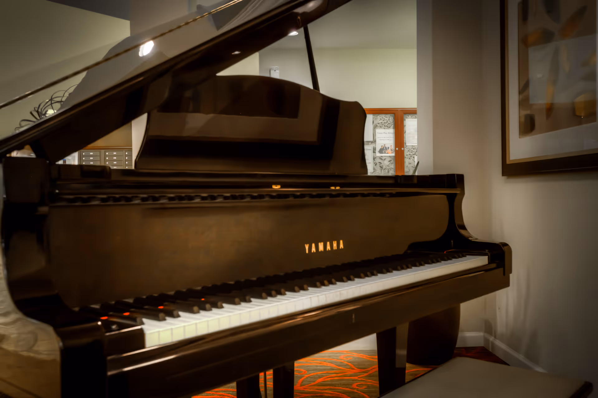 A black Yamaha grand piano in a room with beige walls, a framed abstract artwork on the right wall, and a bulletin board with papers in the background.