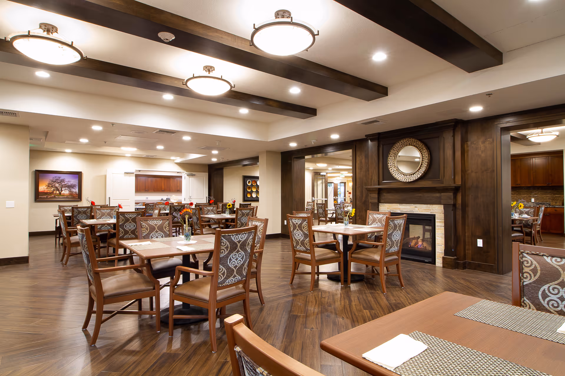 Spacious dining room with multiple wooden tables and upholstered chairs, a fireplace and warm overhead lighting.