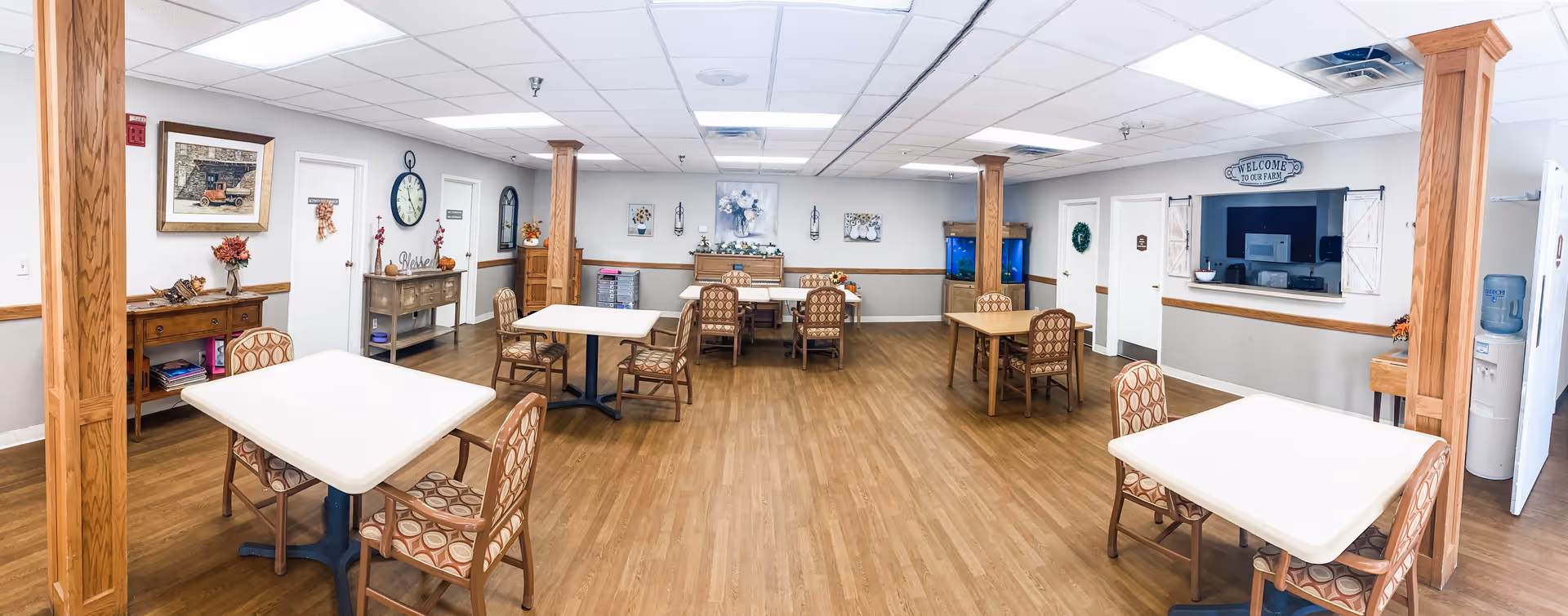 A spacious dining room in a senior living facility with several square tables and patterned chairs arranged neatly on a wood floor. The room features wooden columns, white walls with light brown wainscoting, decorative wall art, a large clock, and a serving window with a 'Welcome to Our Family' sign above it. There is also a water cooler and a small console table with flowers and decor.