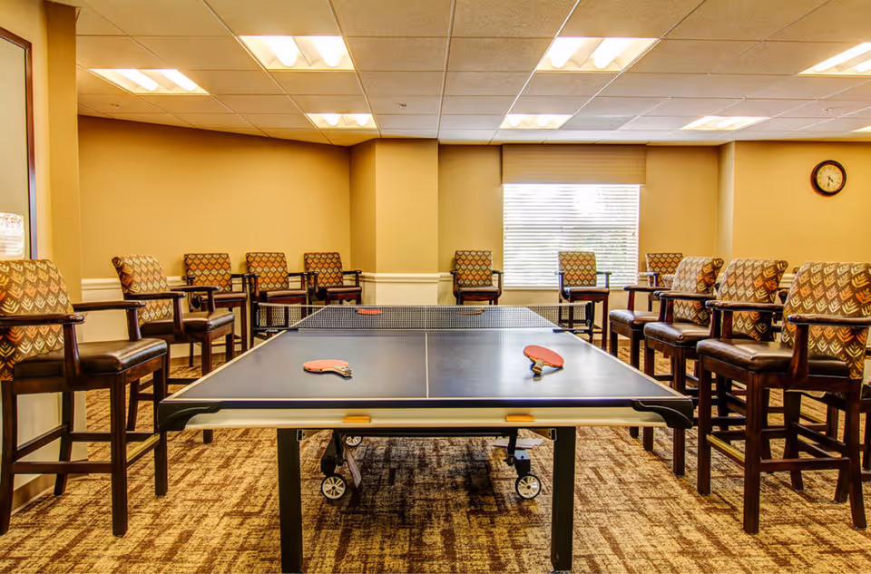 Indoor room with a ping pong table in the center, two paddles resting on the table, surrounded by multiple patterned armchairs arranged along the walls. The room has beige walls, a window with blinds, a clock on the wall, and a carpeted floor.