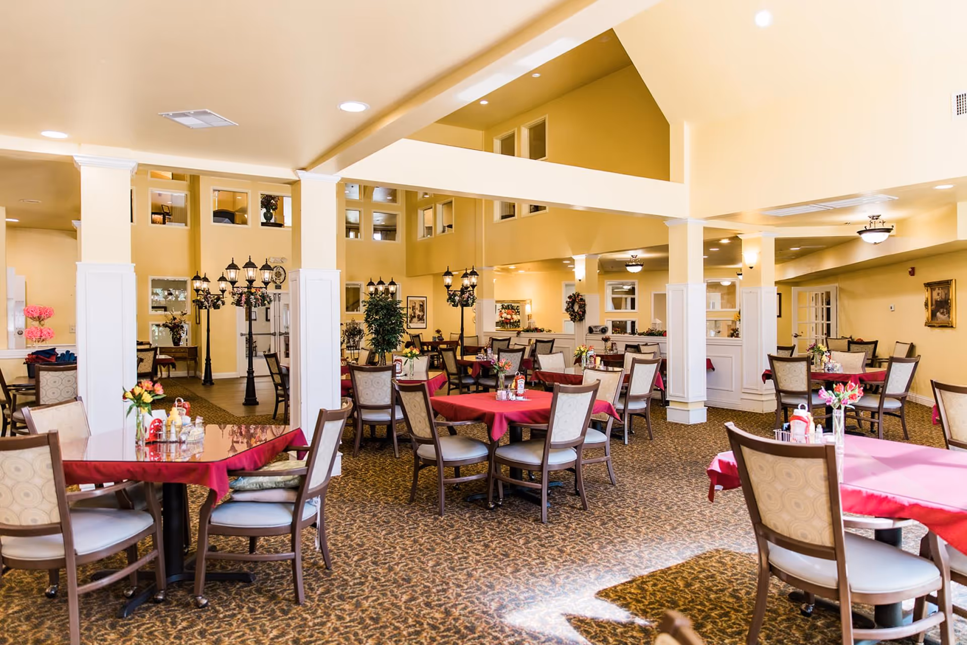 A spacious dining room with multiple tables covered in red tablecloths, each surrounded by cushioned chairs. The room features beige walls, carpeted floors, decorative plants, and vintage-style street lamps. The ceiling is high with recessed lighting and there are windows on the upper walls.