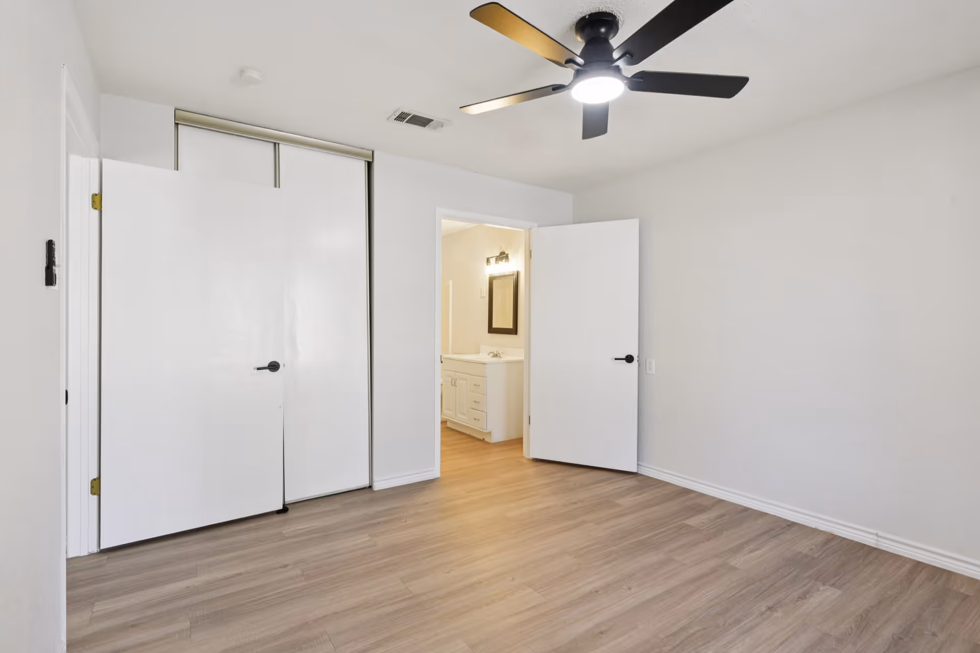 Empty bedroom with light wood flooring, white walls, ceiling fan, closet doors, and adjoining bathroom.