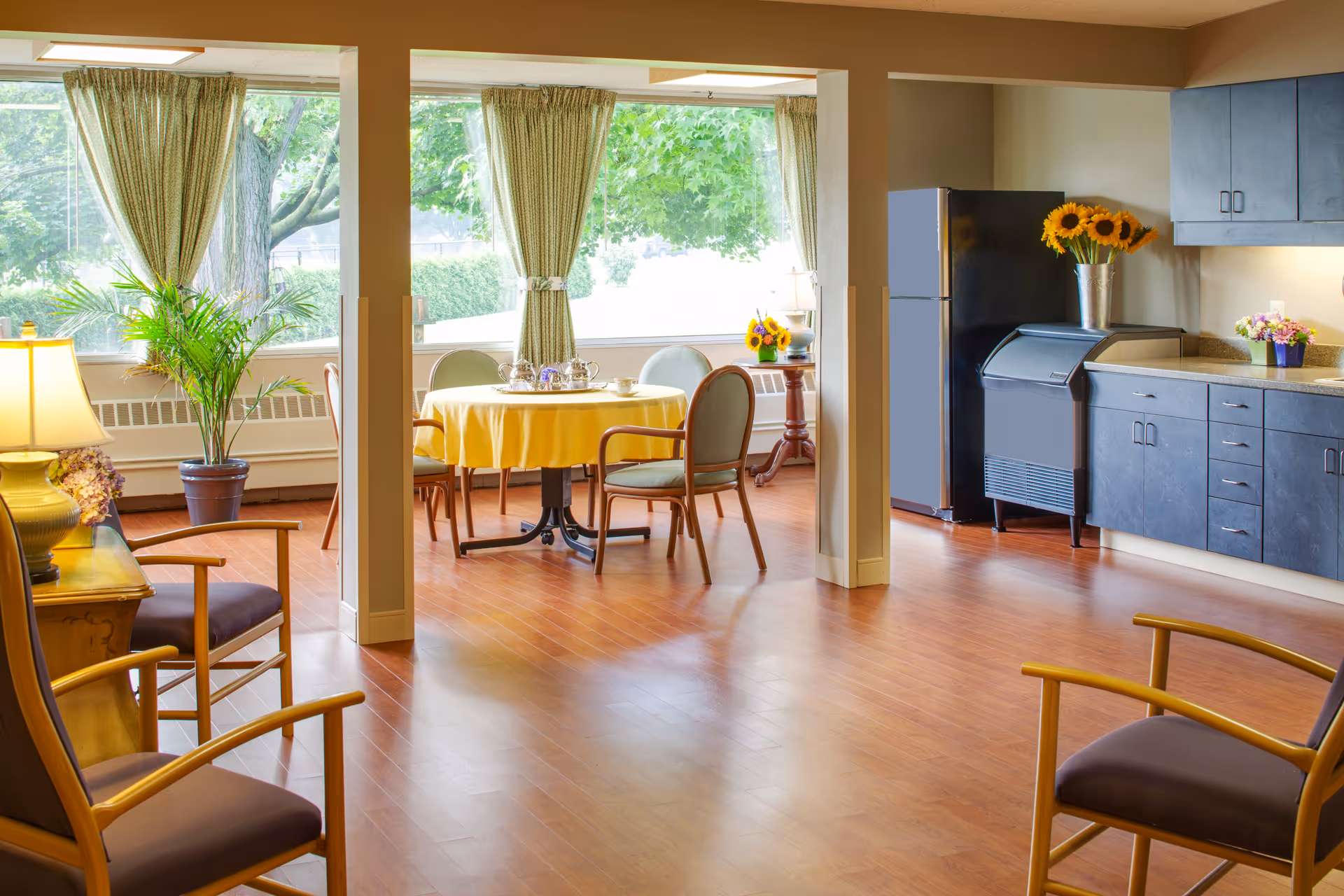 Sunlit communal dining and lounge area with a round table covered in a yellow tablecloth, chairs, a kitchenette, and large windows overlooking trees.