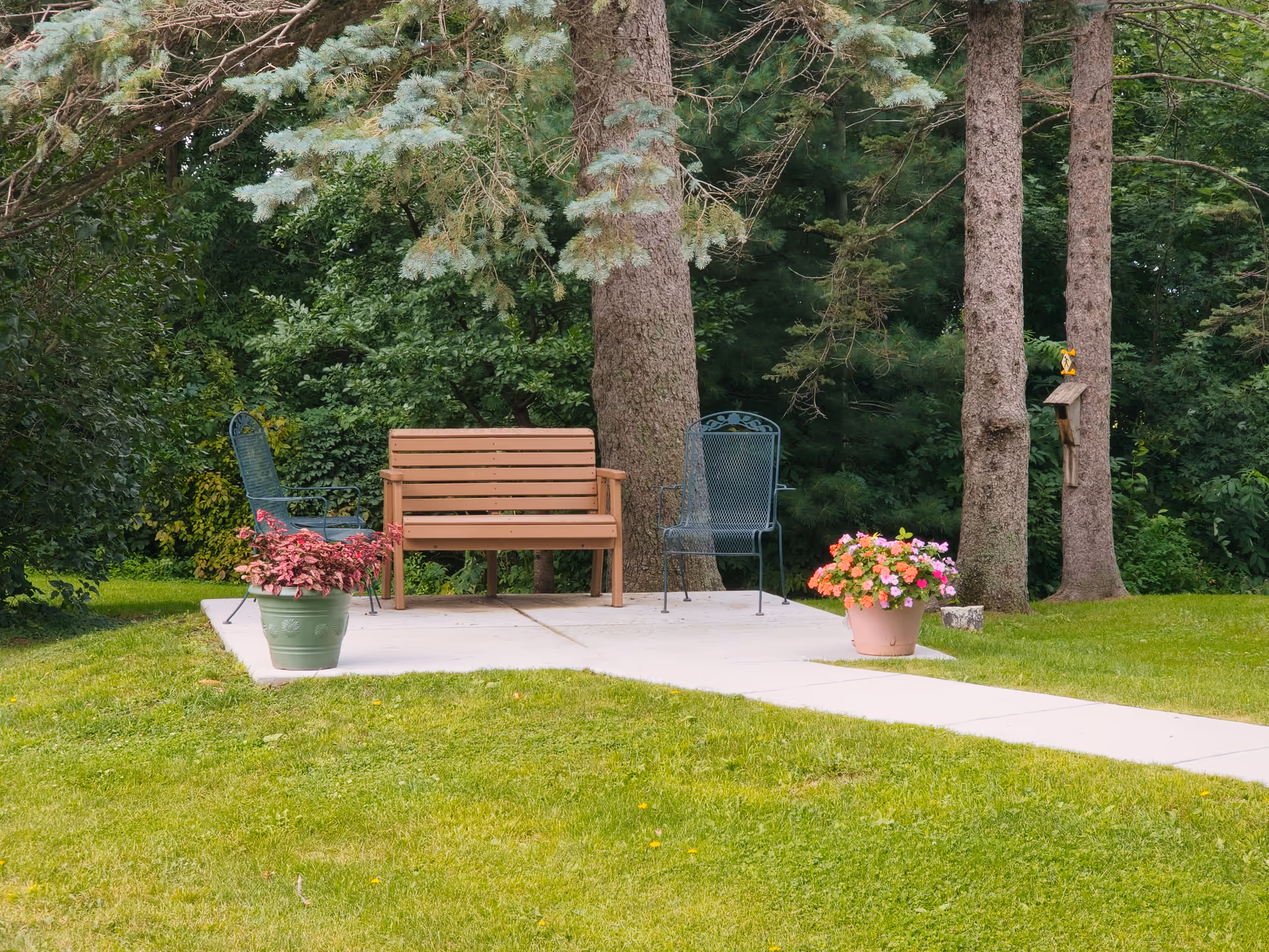 A small outdoor seating area with a wooden bench, two metal chairs, potted flowers, and tall trees in the background.