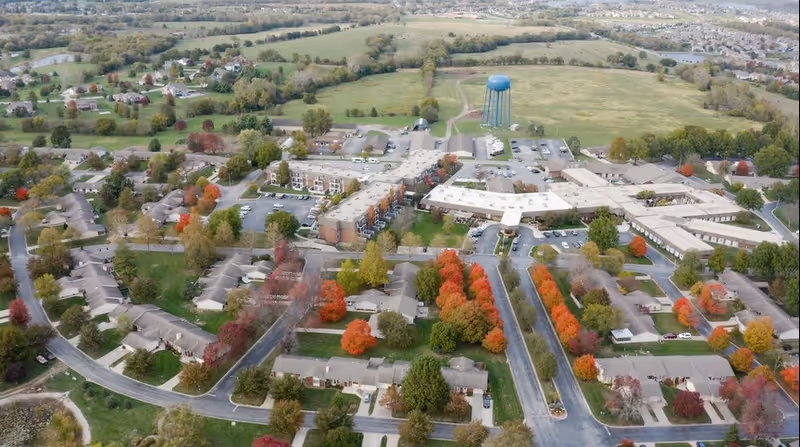 Aerial view of the Foxwood Springs Senior Living campus and surrounding neighborhood with multiple buildings, parking areas, roads, and autumn-colored trees.