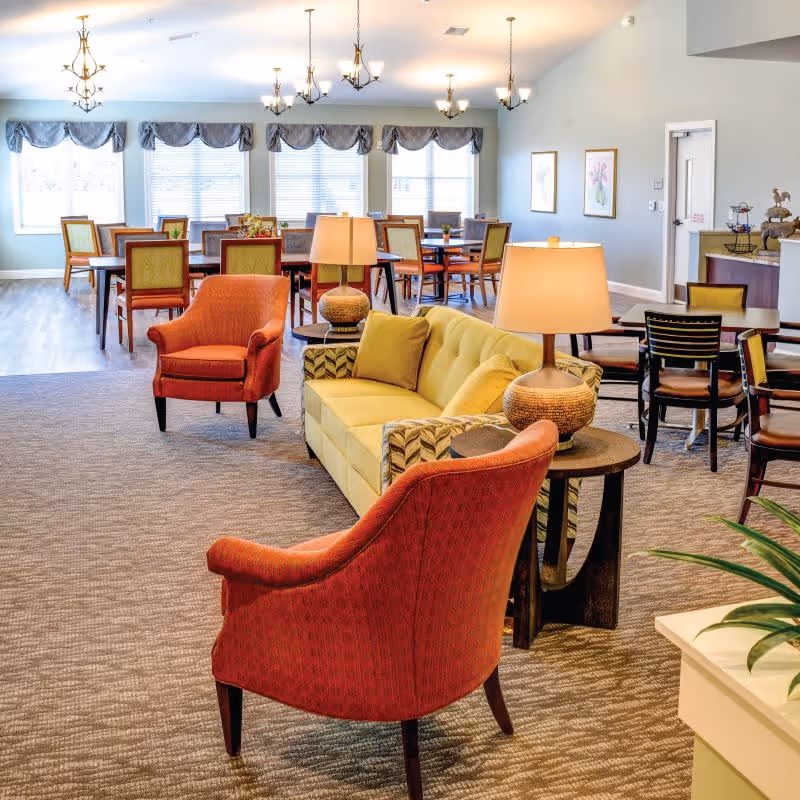A bright and spacious common area in a senior living facility featuring a yellow sofa, two red armchairs, side tables with lamps, and multiple dining tables and chairs near large windows with valances.