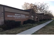 Exterior front view of a single-story brick health and rehabilitation center with a sidewalk, lawn, and trees.