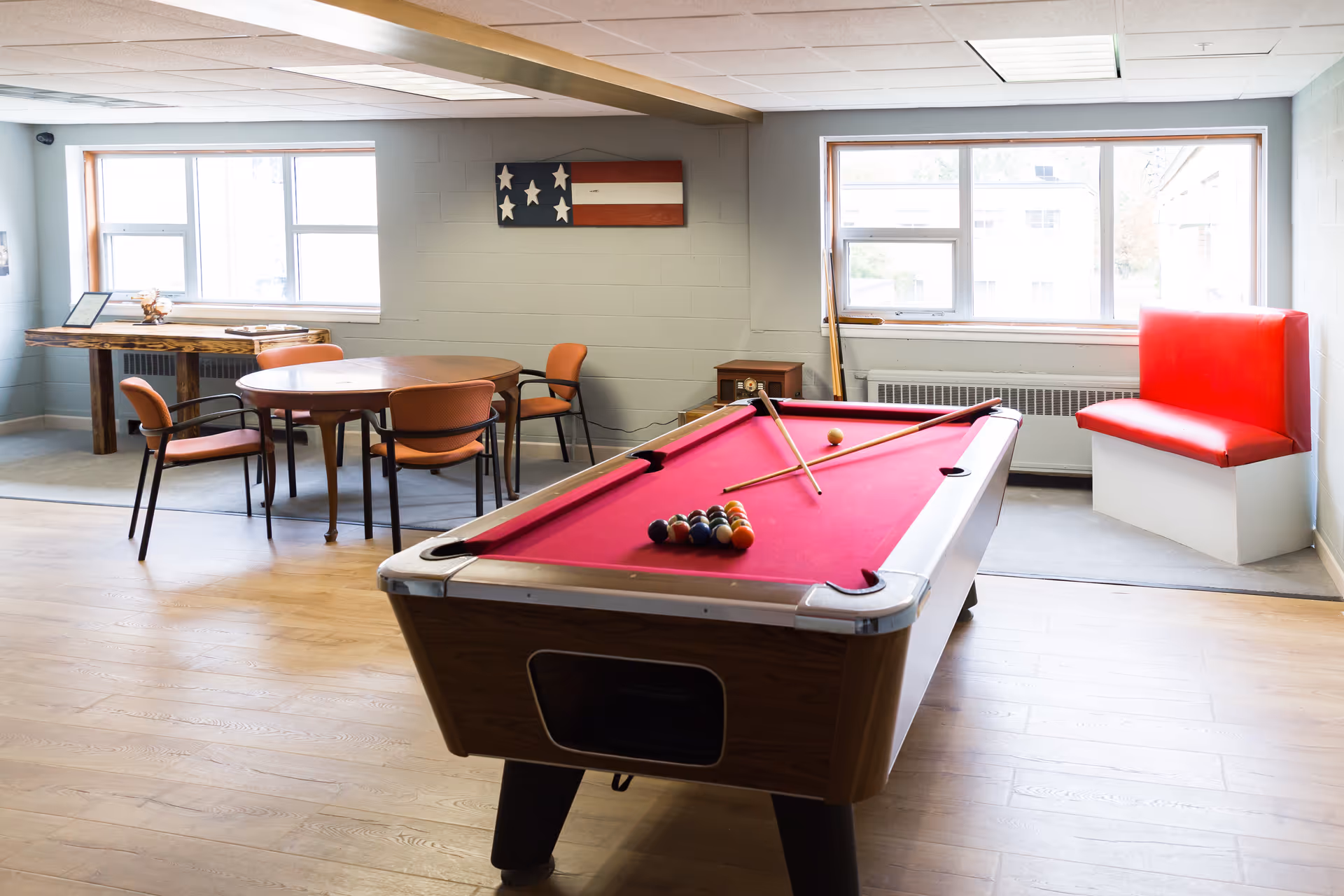A bright recreation room with a red felt pool table in the center, two pool cues resting on it, and a rack of billiard balls arranged. Behind the pool table, there is a round wooden table with four orange chairs. On the wall above the table, there is a decorative wooden American flag. To the right, a red cushioned bench is positioned under a large window letting in natural light. The room has light wood flooring and light gray walls.
