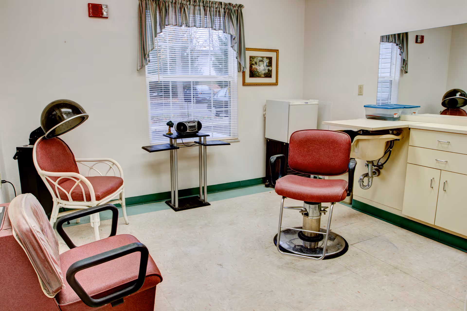 Small salon-style room with red styling chairs, a hooded hair dryer, a sink and cabinets beside a window.