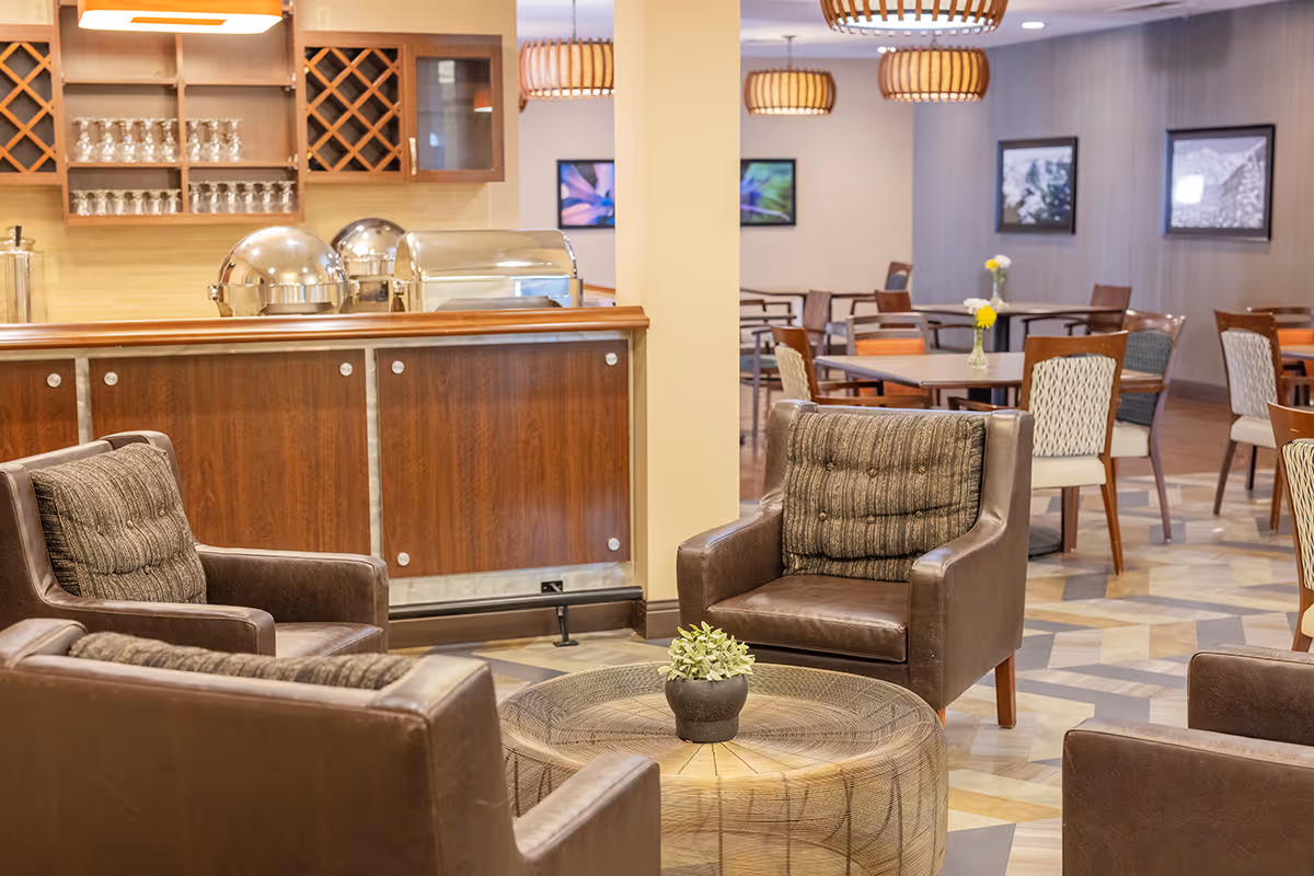 Indoor senior living lounge with brown leather chairs around a round wicker coffee table and a beverage counter in the background.