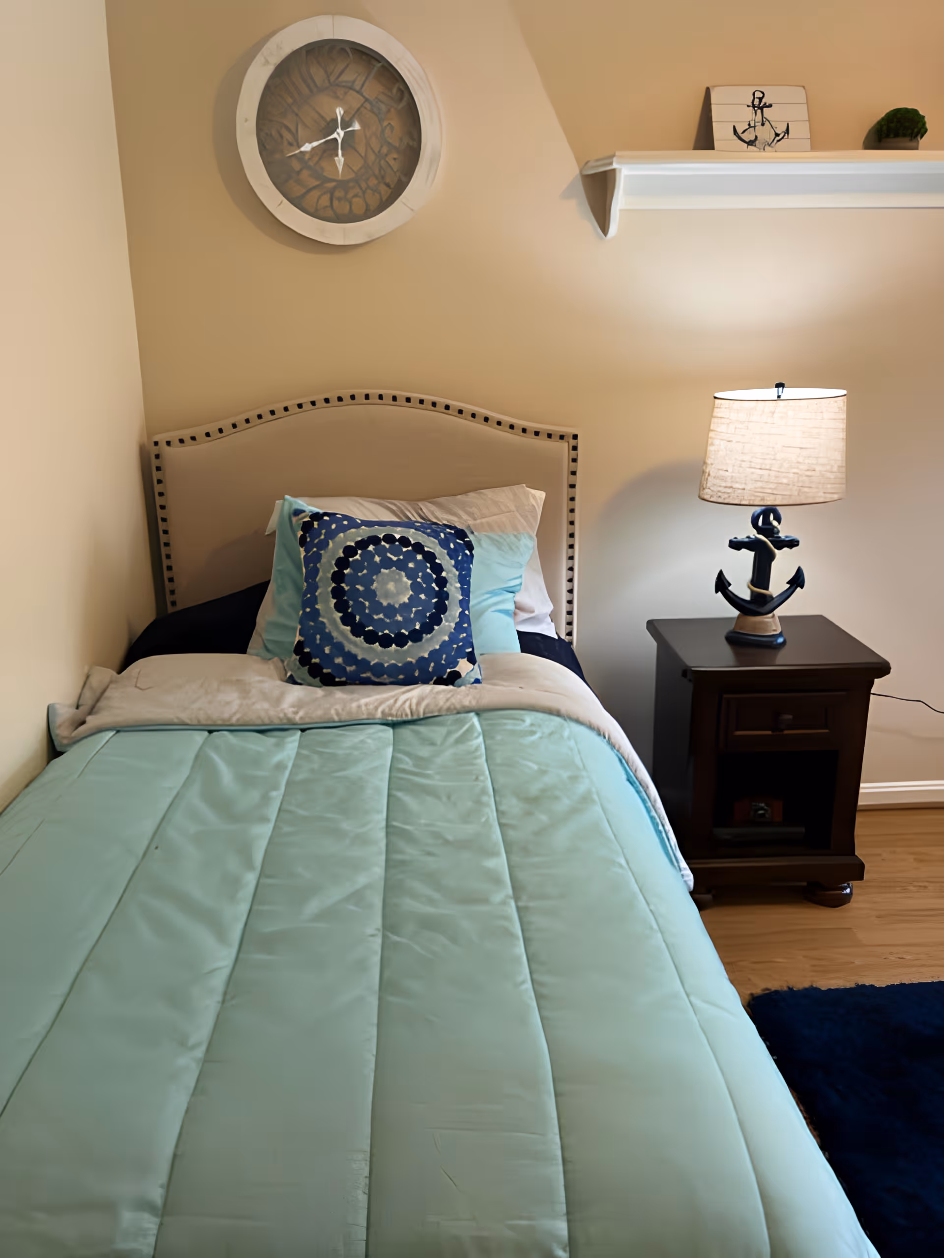 A neatly made single bed with a light blue comforter, multiple pillows including a decorative blue and white circular patterned pillow, and a beige headboard with nailhead trim. On the wall above the bed is a round clock with a rustic design. To the right of the bed is a dark wooden nightstand with a lamp featuring an anchor-shaped base, and a white shelf above it holding a small anchor decoration and a small green plant. The room has beige walls and wooden flooring.