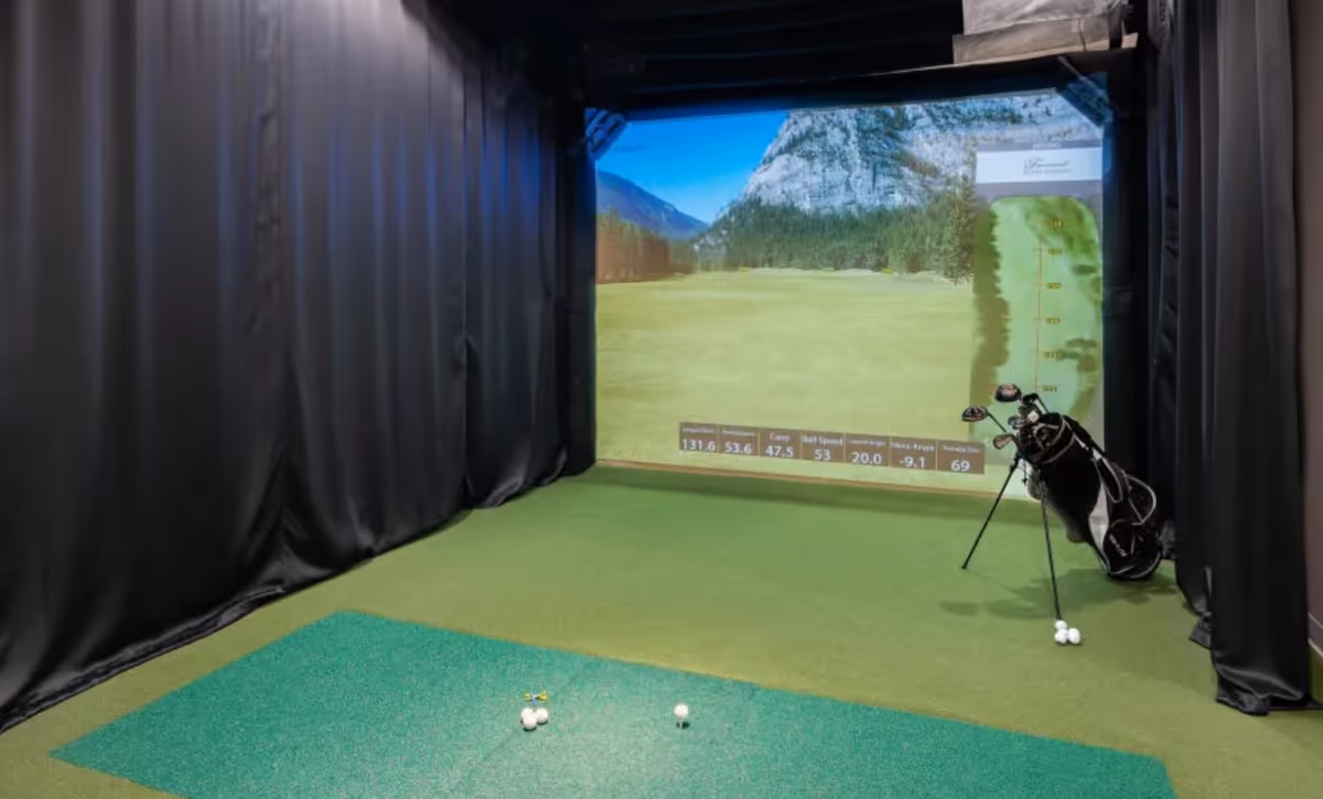 Indoor golf simulator room with a green artificial turf floor, a golf bag with clubs on the right side, and a large screen displaying a golf course scene with various golf statistics.
