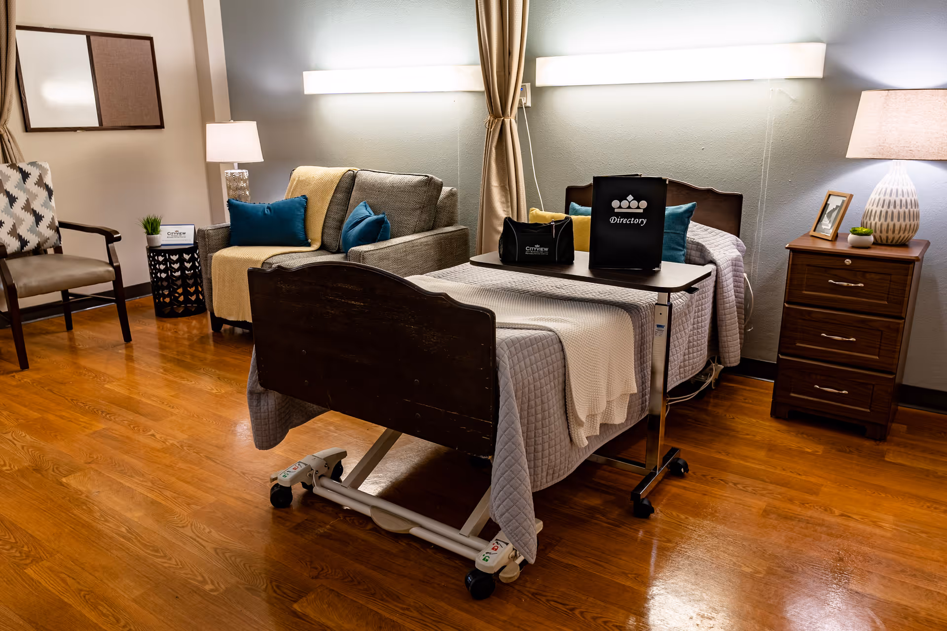 A furnished nursing facility bedroom with a hospital-style bed, overbed table, sofa and nightstand.
