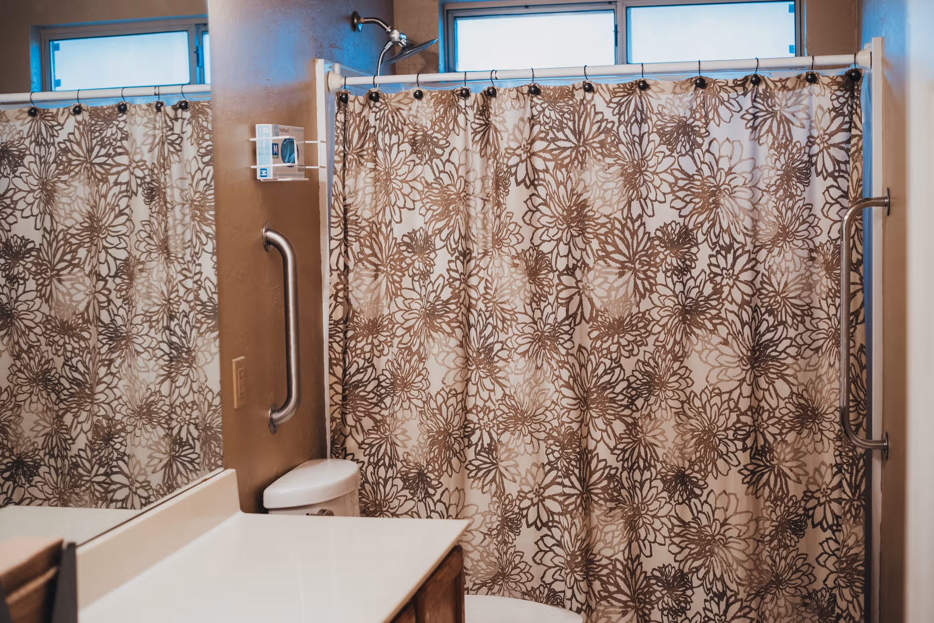 Bathroom with a floral patterned shower curtain, a grab bar on the wall, a toilet partially visible, and a countertop with a mirror reflecting part of the shower curtain.