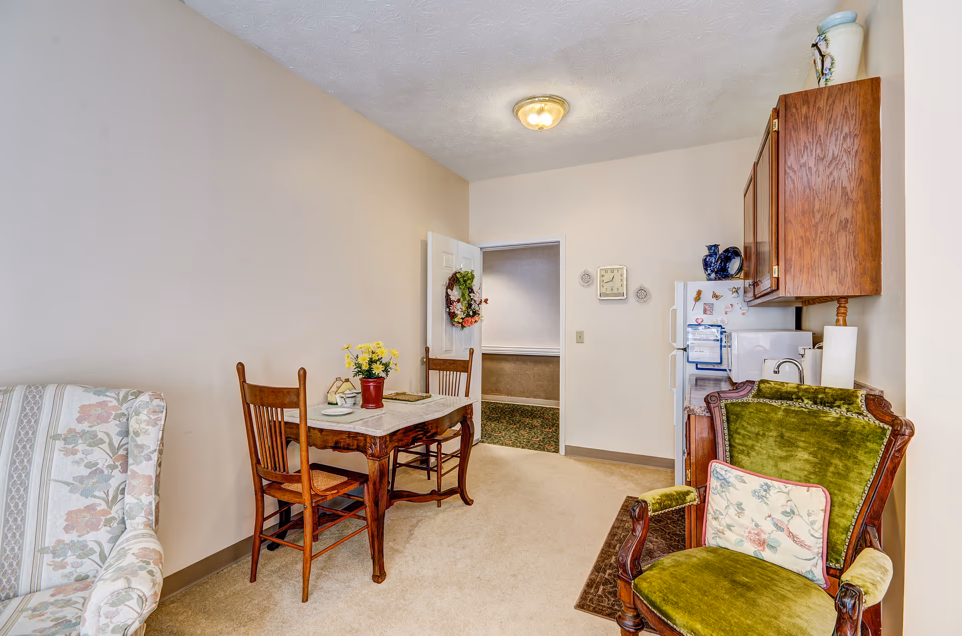 A small dining area in a senior living facility with a wooden table and two wooden chairs. A vase with yellow flowers is on the table. To the right, there is a green upholstered armchair with a floral cushion. In the background, a white refrigerator and wooden cabinets are visible. The walls are light beige, and there is a door leading to another room.