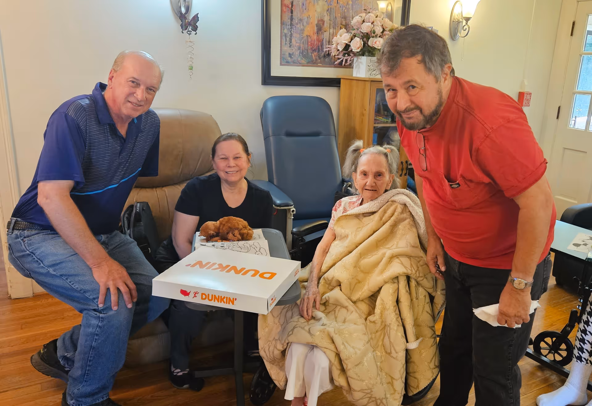 Four people gathered in a cozy living room setting. An elderly woman wrapped in a beige blanket sits in a wheelchair, smiling. Two men and one woman are standing or sitting close by, smiling at the camera. A Dunkin' box and a small brown stuffed animal are on a table in front of them. The room has wooden floors, a beige recliner, a blue chair, a cabinet with flowers, and a door with glass panels.