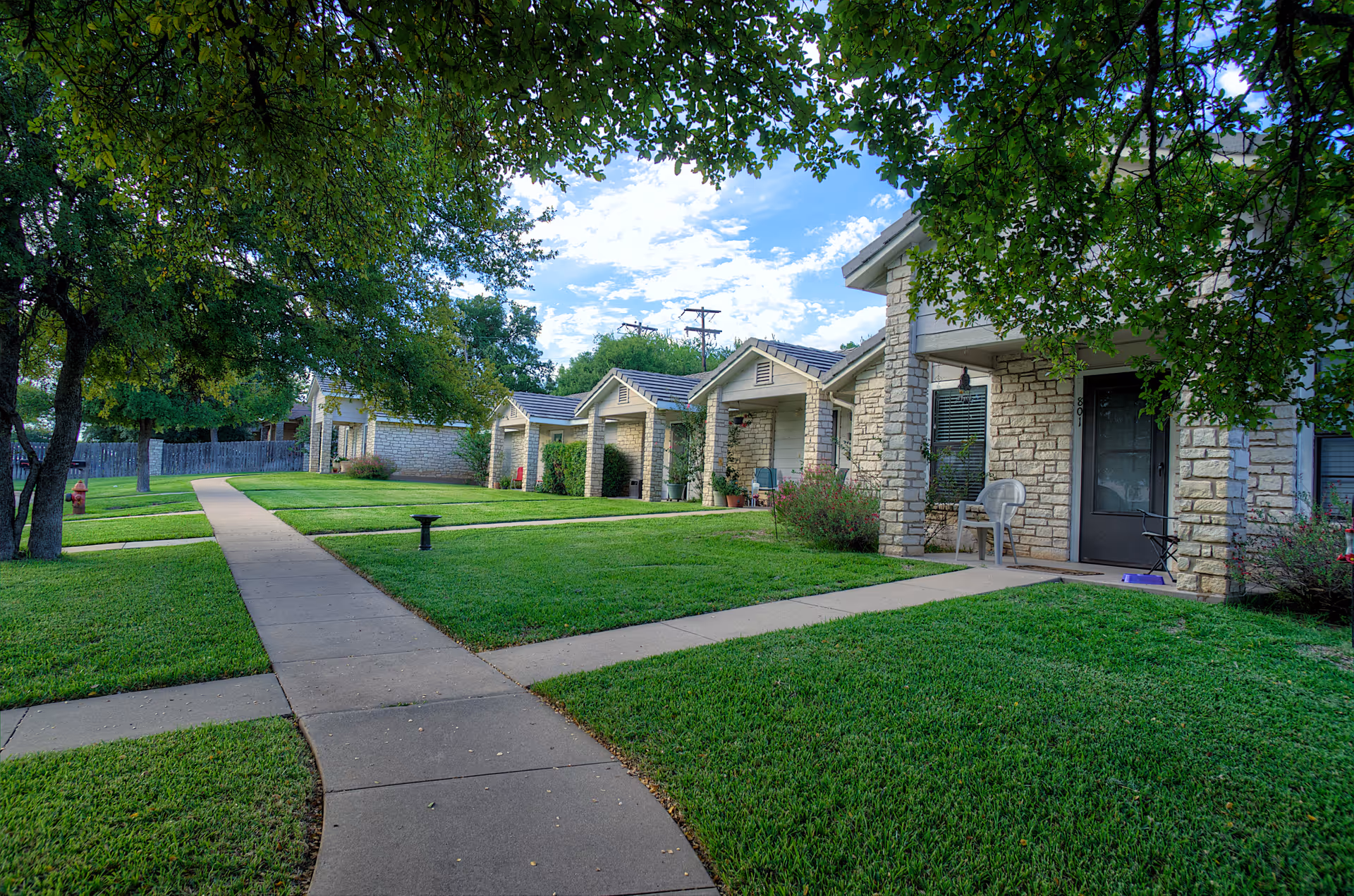 Low-rise stone-faced assisted living units with covered porches, a paved walkway, and well-kept lawns under trees.