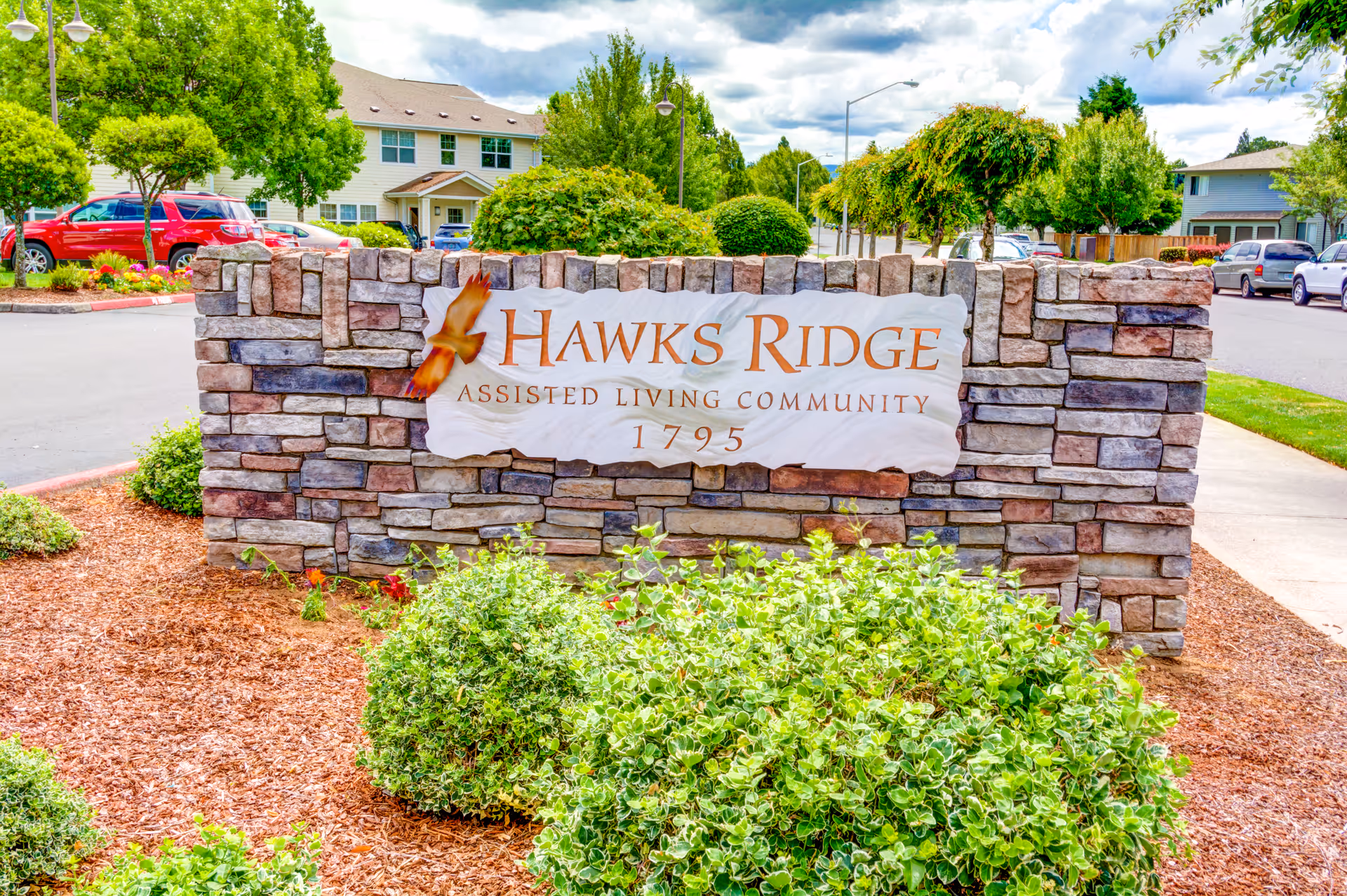 Stone sign for Hawks Ridge Assisted Living Community with the number 1795, surrounded by green bushes and trees, with residential buildings and parked cars in the background under a partly cloudy sky.